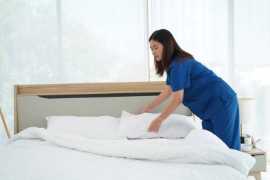 Üniformalı genç bakıcı kadın yatak odasındaki yastıkları ve beyaz yorganı temizliyor ve örtüyordu. Yatma saatine hazırlanıyordu. Yaşlılara bakmak için günlük aktiviteler.