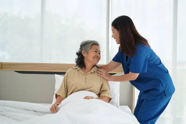 Compassionate nurse assistant offering gentle care to senior woman lying in bed. Young caregiver ensures elderly lady\'s comfort and warmth, covering her with soft blanket for restful sleep at home.