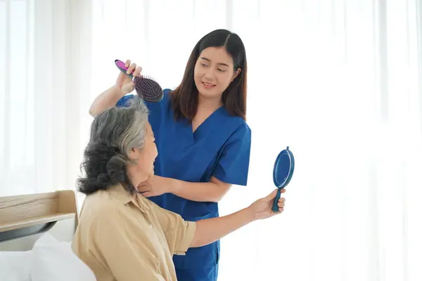 young nurse assistant female gently cares for an elderly woman, gently combing her hair on the bed, Senior woman looking a mirror with happy,  Caregiver providing comfort and reassurance at home