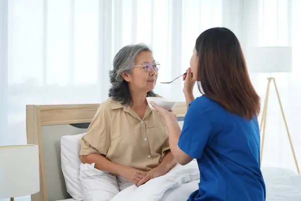 Asian young caregiver female help with feeding breakfast to aged woman on bed, Nurse assistant support and taking care of female patient at home, They are smile and talking with happy together