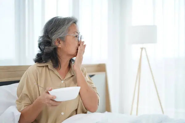 Asian Senior woman wearing glasses sitting on bed in bedroom use hand closed her nose during hold a bowl of meal, expression facial emotion with feeling anorexic