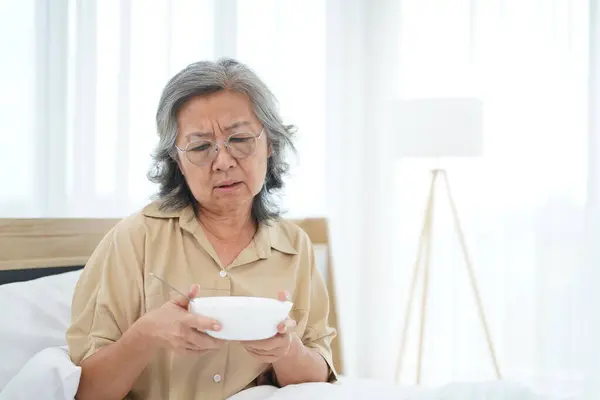 Asian Senior woman wearing glasses sitting on bed in bedroom hold and looking a bowl of meal, expression facial emotion with feeling anorexic
