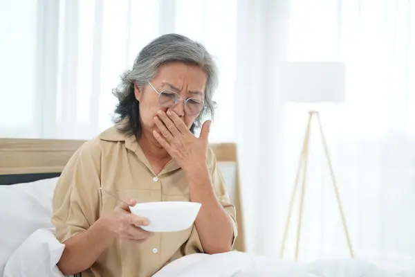 Asian Senior woman wearing glasses sitting on bed in bedroom use hand closed her nose during hold a bowl of meal, expression facial emotion with feeling anorexic