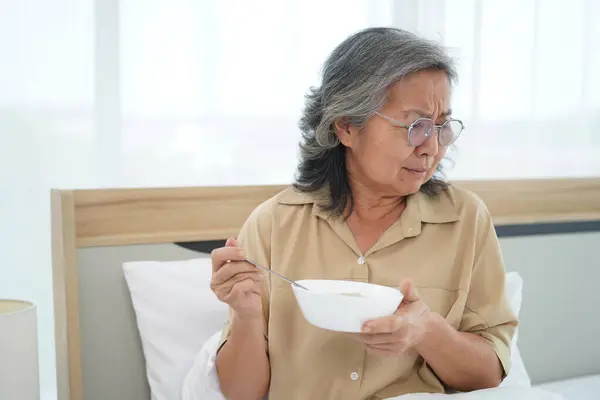 Asian Senior woman wearing glasses sitting on bed in bedroom hold a bowl of meal, expression facial emotion with feeling anorexic