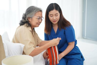 Asyalı son sınıf öğrencisi yatak odasında oturuyor ve hobi olarak el işi ya da iş terapisi olarak el işi tığ işi yapıyor genç kadın bakıcıya öğretmenlik yapıyor evde ya da huzurevinde birlikte eğleniyorlar.