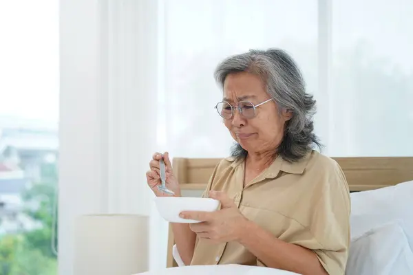 Asian Senior woman wearing glasses sitting on bed in bedroom hold and looking a bowl of meal, expression facial emotion with feeling anorexic and distasteful
