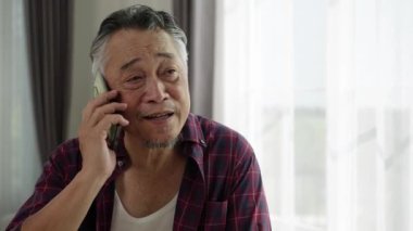 Ekose gömlekli gülümseyen yaşlı Asyalı adamın parlak pencerenin önünde akıllı telefondan konuşmasının el kamerasıyla çekilmiş videosu. Doğal ışık onun dost ifadesini aydınlatır. Rastgele ev ayarlama sıcaklığı ve bağlantıyı iletir.