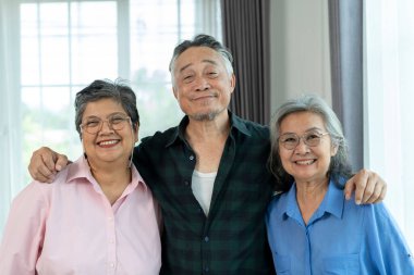 Asyalı olgun bir adamın portresi iki kadın arasında duruyor ve yakın arkadaşlarının omuzlarında kollarıyla sarılıyor, gülümsüyor ve kameraya bakıyor.