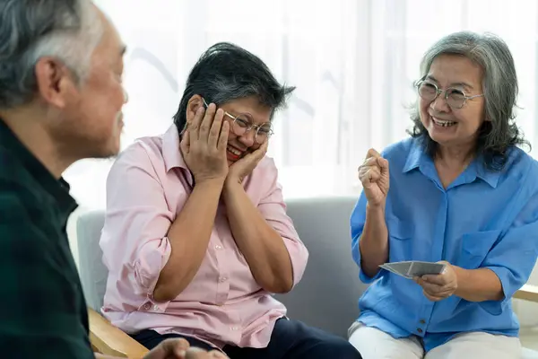 stock image Selective focus, Senior Asian woman holds cards, hoping to win the game, and two friends cheer her on. Cheerful retired Asian friends spending time together with happy in comfortable living room