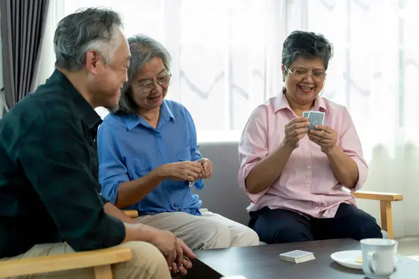 stock image Selective focus, senior Asian friends during retirement. A woman with playing cards converses with a man and another woman in a bright, cozy living room, creating a casual and relaxed atmosphere