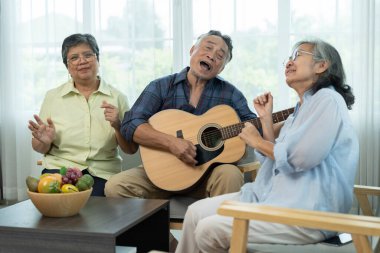 Üç Asyalı mutlu yaşlı arkadaş müzik ve arkadaşlığın tadını çıkarıyor, olgun bir adam akustik gitar tıngırdatırken, bayan arkadaşlar parlak bir oturma odasında şarkı söylüyorlar..