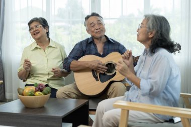 Üç Asyalı mutlu yaşlı arkadaş müzik ve arkadaşlığın tadını çıkarıyor, olgun bir adam akustik gitar tıngırdatırken, bayan arkadaşlar parlak bir oturma odasında şarkı söylüyorlar..