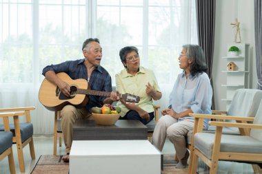 Üç Asyalı mutlu yaşlı arkadaş müzik ve arkadaşlığın tadını çıkarıyor, olgun bir adam akustik gitar tıngırdatırken, bayan arkadaşlar parlak bir oturma odasında şarkı söylüyorlar..