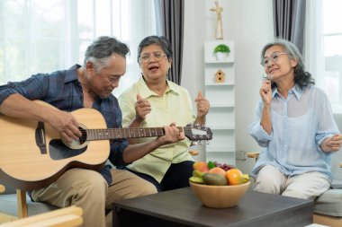 Üç Asyalı mutlu yaşlı arkadaş müzik ve arkadaşlığın tadını çıkarıyor, olgun bir adam akustik gitar tıngırdatırken, bayan arkadaşlar parlak bir oturma odasında şarkı söylüyorlar..