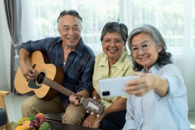 Seçici odaklanma, üç mutlu kıdemli arkadaş kapalı bir yerde toplandı, biri akustik gitar çalarken diğerleri animasyonlu bir şekilde şarkı söylüyor, bayan arkadaş oturma odasında arkadaşlığı ile akıllı telefon selfie 'leri kullanıyor.