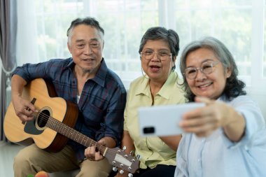 Seçici odaklanma, üç mutlu kıdemli arkadaş kapalı bir yerde toplandı, biri akustik gitar çalarken diğerleri animasyonlu bir şekilde şarkı söylüyor, bayan arkadaş oturma odasında arkadaşlığı ile akıllı telefon selfie 'leri kullanıyor.