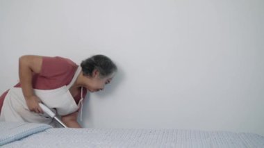 Önlük takan bir ev hanımı yatak odasını paspasla temizliyor. Alanın lekesiz ve bakımlı olmasını sağlıyor. Temizliğe olan bağlılığı çok açık..