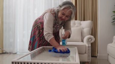 Ev hanımı renkli bir elbise içinde, mavi bir bez ve temizlik spreyiyle dikkatle cam bir sehpayı temizliyor..