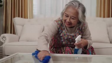Ev hanımı renkli bir elbise içinde, mavi bir bez ve temizlik spreyiyle dikkatle cam bir sehpayı temizliyor..