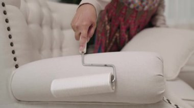 Yakın plan çekim görüntüsü, renkli elbiseli hizmetçi oturma odasındaki beyaz koltuğu temizlemek için iplik rulo kullanıyor, lekesiz kalmasını sağlıyor..