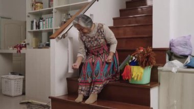 El kamerası görüntüsü, yaşlı bir temizlikçi merdivenlerde oturur, acı dolu bir ifadeyle sırtını sıvazlar, temizlik malzemeleriyle çevrilidir. Yorgun görünüyor ve diz ve sırt ağrısı çekiyor..