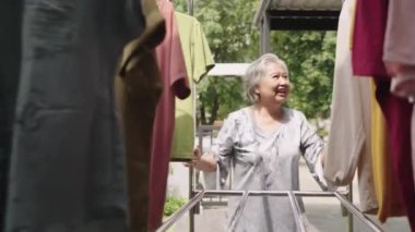 Orta büyüklükte gülümseyen Asyalı bir kadın giysiler kuruduktan sonra çamaşır ipini arka bahçeden itiyor. Yaşlı bir kadın çamaşırhanede rutin bir iş yapıyor.