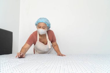 Koruyucu maske takan orta yaşlı bir temizlikçi yatak odasındaki çarşafları serer. Temizlik ve düzenli bir çevreyi korumaya olan bağlılığını gösterir..