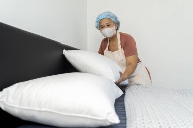 Asyalı temizlikçi önlük takar, yastık getirir, yatağa koyar, yatağın düzenini düzenler ve kontrol eder. Ayrıntılara ve düzenliliğe dikkat et, boşluğu kopyala