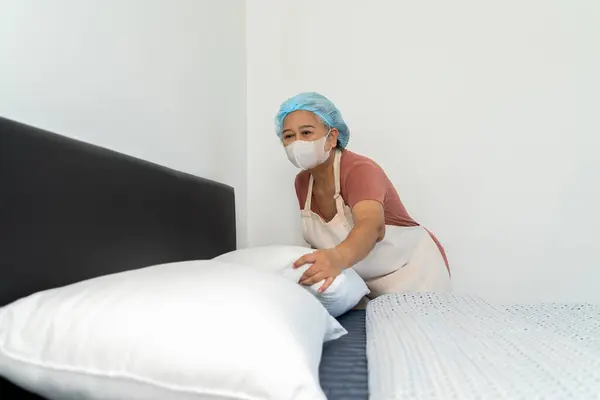 stock image Asian housekeeper wears an apron, brings in pillows, puts them on the bed, organizes and checks the neatness of the bed. Attention to detail and orderliness, copy space