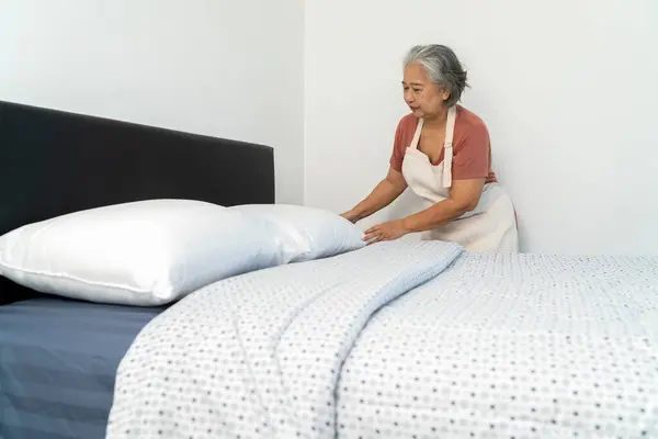 stock image Asian housekeeper wears an apron, brings in pillows, puts them on the bed, organizes and checks the neatness of the bed. Attention to detail and orderliness, copy space