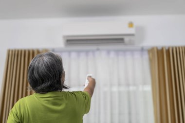 Arka görüş, Asyalı Yaşlı Kadın Tutma ve modern oturma odasındaki klimayı ayarlamak için uzaktan kumanda kullanıyor