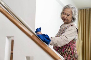 Asian mature woman in colorful dress cleaning stair railing with a cloth, housemaid maintaining cleanliness at home.