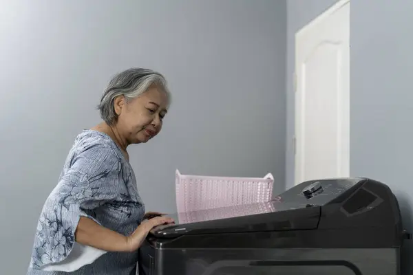 stock image Asian Senior Woman Laundry clothes and touch on button to operate a washing machine in laundry room, housemaid focusing on her household chores.