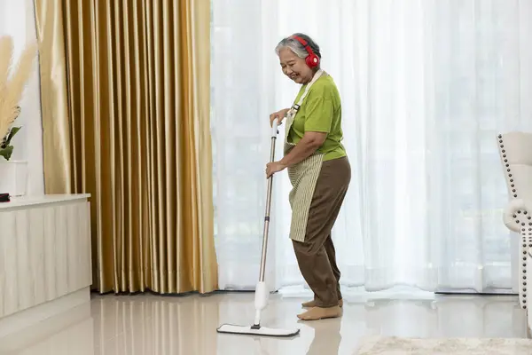 stock image Portrait of Asian middle aged female housekeeper in apron and headphones joyfully dancing while mopping the floor in living room, copy space