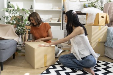 İki genç Asyalı kadın yaşam alanına karton kutuları neşeyle açıyor, bitkiler ve doğal ışıkla çevrili, dostluk ve keşif anı sergiliyorlar, birlikte konuşuyorlar ve mutlu bir şekilde gülümsüyorlar.