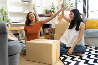 İki genç Asyalı kadın rahat oturma odasında yerde oturup birbirlerine beşlik çakıyor ve ev dekoru ve eşyalarıyla çevrili kutuları başarıyla taşıdıktan sonra mutlu bir şekilde gülümsüyor..