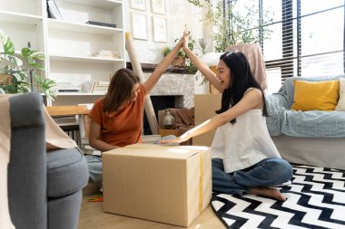 İki genç Asyalı kadın rahat oturma odasında yerde oturup birbirlerine beşlik çakıyor ve ev dekoru ve eşyalarıyla çevrili kutuları başarıyla taşıdıktan sonra mutlu bir şekilde gülümsüyor..