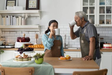 Asyalı, gülümseyen son sınıf çifti modern bir mutfakta kahvelerini yudumlarken ellerinde sıcak bir içecek tutuyorlar ve birlikte konuşmaktan zevk alıyorlar. Olgun çiftler evlerinde mutluluğu paylaşırlar.