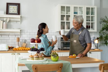 Önlüklü yaşlı çift, etrafları hamur işleri ve meyvelerle çevrili, rahat bir mutfakta kahvelerini paylaşıyor, huzurlu bir sabahın tadını çıkarıyorlar, olgun çiftler evlerinde mutluluklarını paylaşıyorlar.