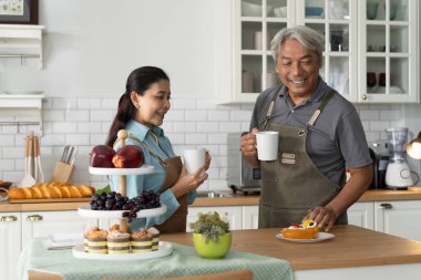 Önlüklü yaşlı çift, etrafları hamur işleri ve meyvelerle çevrili, rahat bir mutfakta kahvelerini paylaşıyor, huzurlu bir sabahın tadını çıkarıyorlar, olgun çiftler evlerinde mutluluklarını paylaşıyorlar.