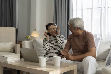 Kulaklık takan Asyalı güzel bir kadın, rahat bir oturma odasında yaşlı bir adamla müzik dinlemekten hoşlanıyor, karı koca neşeli bir şekilde akıllı telefondan şarkı seçiyor, evde birlikte mutluluğu paylaşıyorlar.