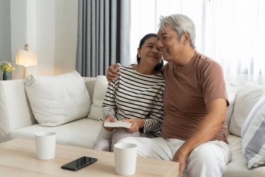 Asyalı yaşlı çift kanepede oturuyor ve oturma odasında mutlu bir gülümsemeyle kitap okuyor, konuşurken mutlulukla kucaklaşıyorlar, neşeli çift evde birlikte vakit geçiriyorlar.
