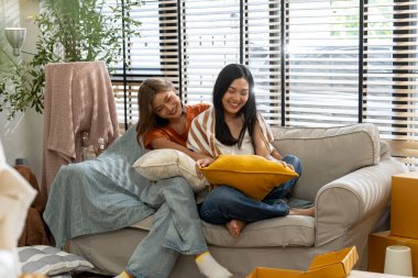 İki genç Asyalı kadın, parlak, aydınlık bir oturma odasındaki kutuları taşıyan rahat bir kanepede sarılıp gülüyorlar. Mutlu lezbiyen çift taşınma gününden sonra yeni evlerinde dinleniyor.