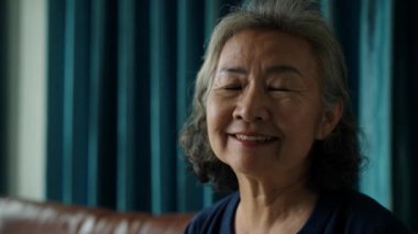 Kilitli çekim, gülümseyen Asyalı son sınıf öğrencisinin yüzüne yakın plan portre, yüzünü kameraya ve dişlek gülümsemeye çevir, mavi perdenin önüne otur, sıcaklığı ve pozitifliği ifade et.