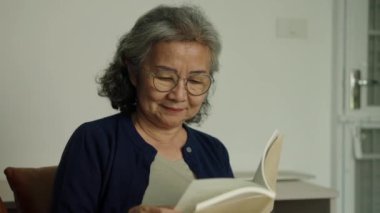 Dolly orta boy, gözlüklü yaşlı kadının kanepede oturduğu ve rahat ev ortamıyla çevrili kitap okumaktan zevk aldığı görüntüler, evde dinlenirken eğlenerek gülümseyip güler.