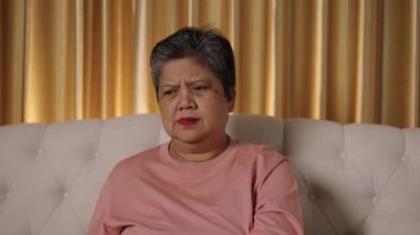 Görüntüler orta ölçekli, Asya 'lı yaşlı bir kadının kısa saçlı yüzü oturma odasında kanepede otururken rahatsız edici, düşünceli ve tatminsiz bir ifade sergiliyor.