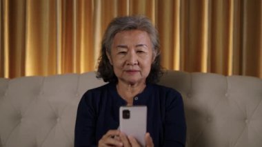 Görüntüler orta ölçekli, Asyalı yaşlı bir kadın koltukta oturuyor, akıllı telefonuna odaklanmış bir ifadeyle bakıyor, muhtemelen bakınıyor ya da mesajlaşıyor..