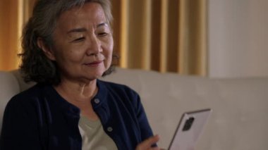 Görüntüler kilitli, koltukta oturan Asyalı yaşlı kadının yüzünü kapat, akıllı telefonuna odaklanmış bir ifadeyle bak, muhtemelen bakınıyor ya da mesaj atıyor..