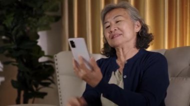 Görüntüler kilitli, neşeli Asyalı son sınıf kadını oturma odasında akıllı telefonuyla dinleniyor, müzik dinliyor ve cep telefonuna mutlulukla bakıp gülümsüyor.