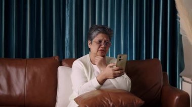 Görüntüler orta ölçekli, Yaşlı Kadın video görüşmesi sırasında akıllı telefonla konuşurken endişeli görünüyor, oturma odasındaki ciddi bir konuşmada endişe verici haberler alıyor veya alıyor